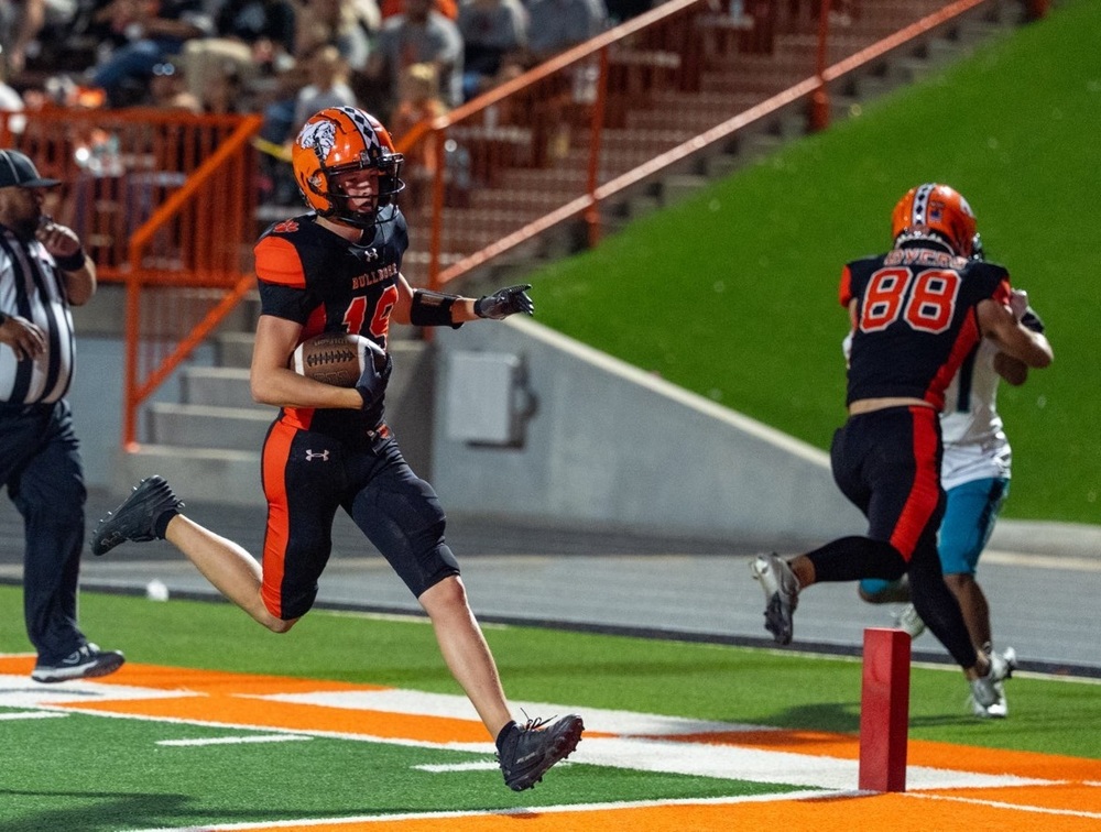 A teen boy in a black-and-orange football uniform runs over the goal line with the football tucked in his right elbow as a teammate blocks an opponent in white and teal.