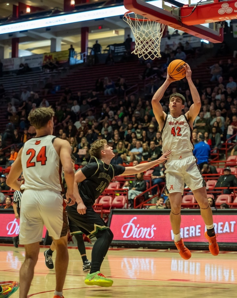 A teen boy in a white and orange basketball uniform holds the ball above his head in both hands as he jumps in preparation for a shot. An opponent in black pushes him with his left hand as a teammate looks on.