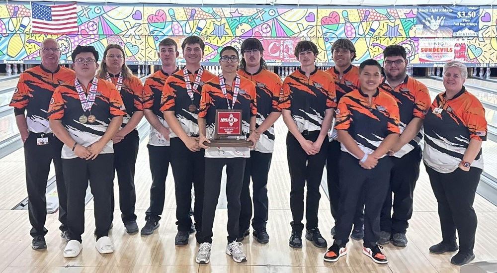 Ten male bowlers, four wearing medals around their necks and one in the center holding a red trophy, pose for a photo in front of bowling lanes with a male coach on the far left and a female coach on the far right.