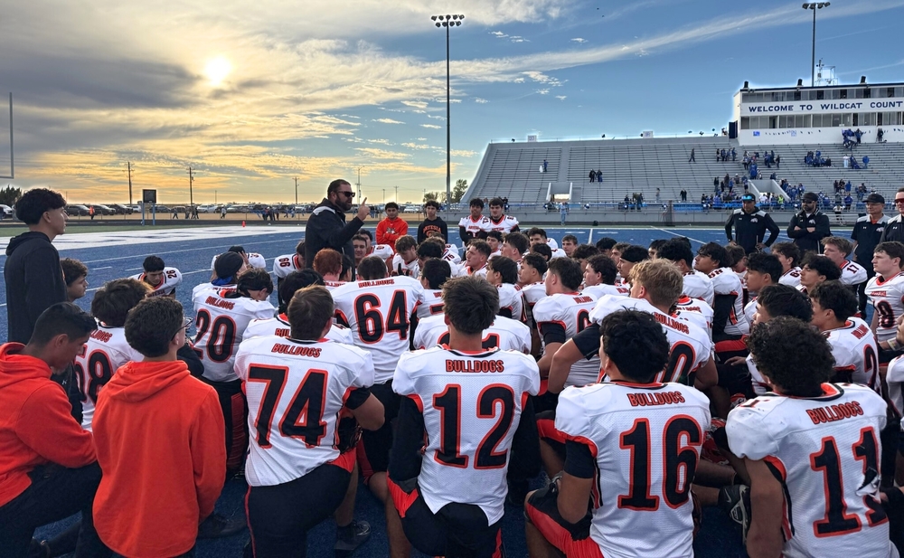 A large group of teen boys in white, orange and black football uniforms kneel with their backs to the camera facing a male coach who is holding his index finger up as he speaks to them. The sun can be seen setting on a blue football field in front of a grandstand in the background.