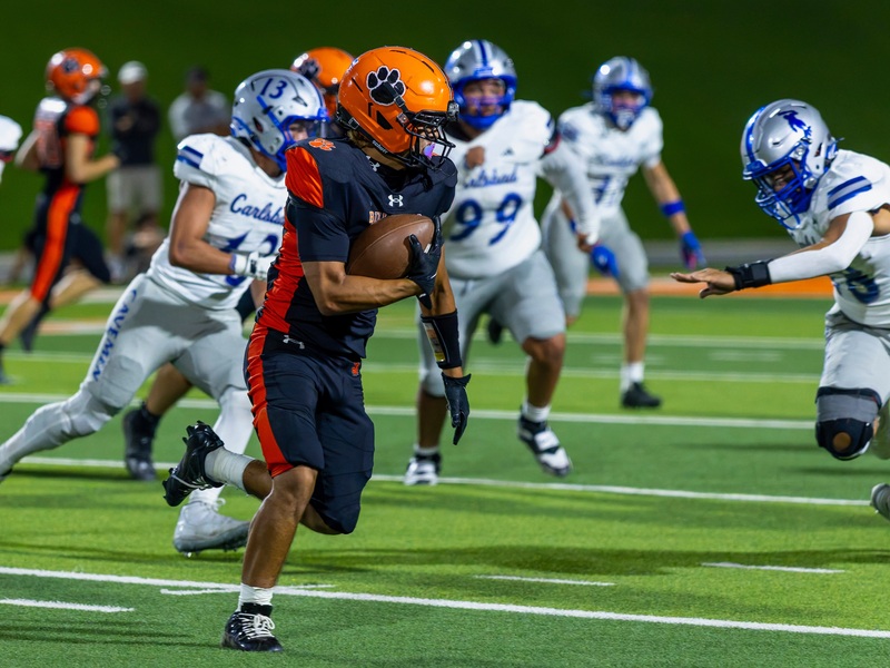 A teen boy in a black and orange football uniform cradles the ball in his right elbow as he runs past a host of defenders in silver and blue.