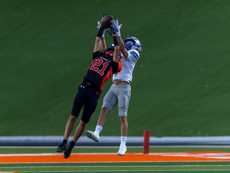 A teen boy in a black and orange football uniform leaps into the air in front of a receiver in silver and blue to break up a pass.