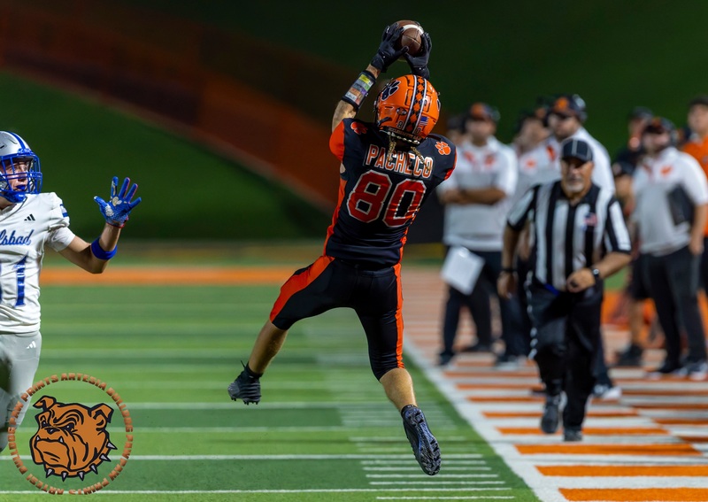 A teen boy in a black and orange football uniform leaps into the air to catch a pass as an opponent in silver and blue looks on.