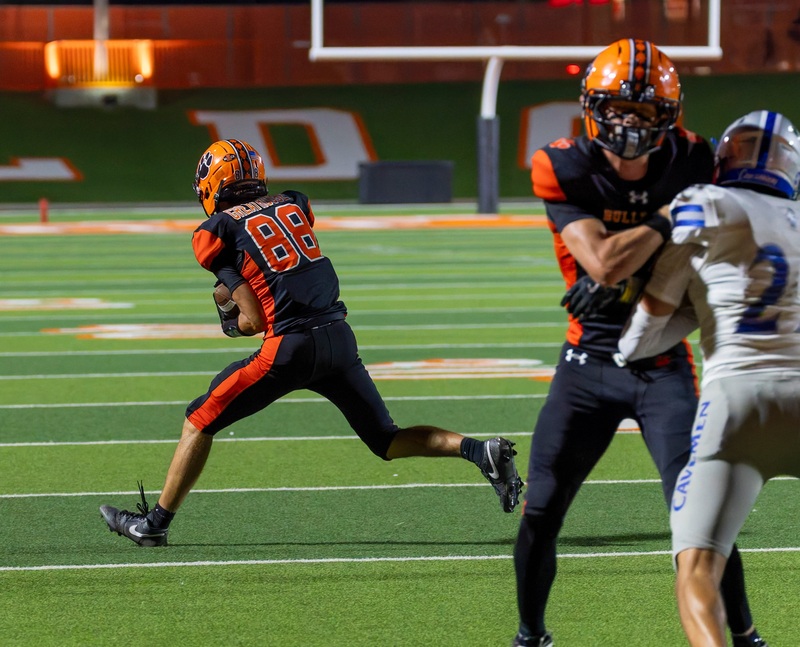 A teen boy in a black and orange football uniform comes down with a catch as a teammate blocks an opponent in silver and blue.