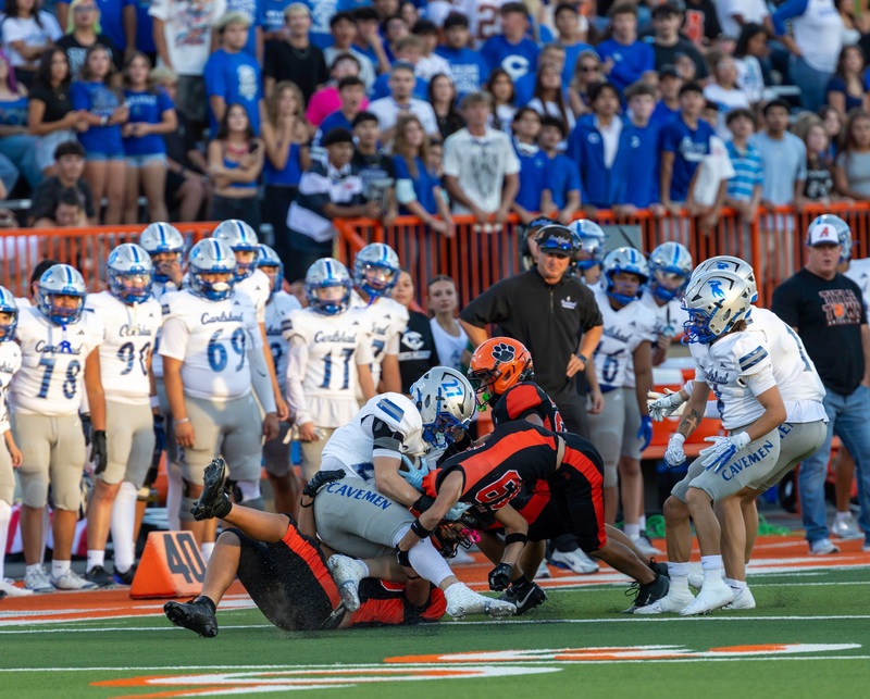 Three teen boys in black and orange football uniforms tackle a running back in silver and blue.