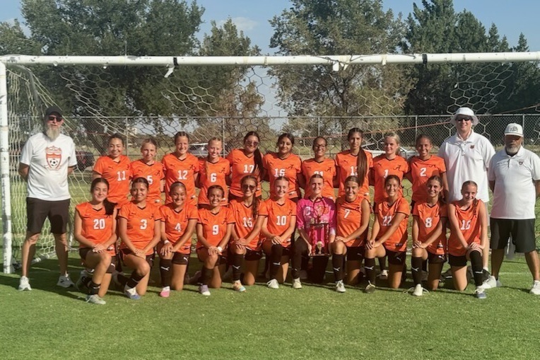 A girls' soccer team in orange uniforms and three male coaches in white and black pose for a photo in front of a goal with a trophy.