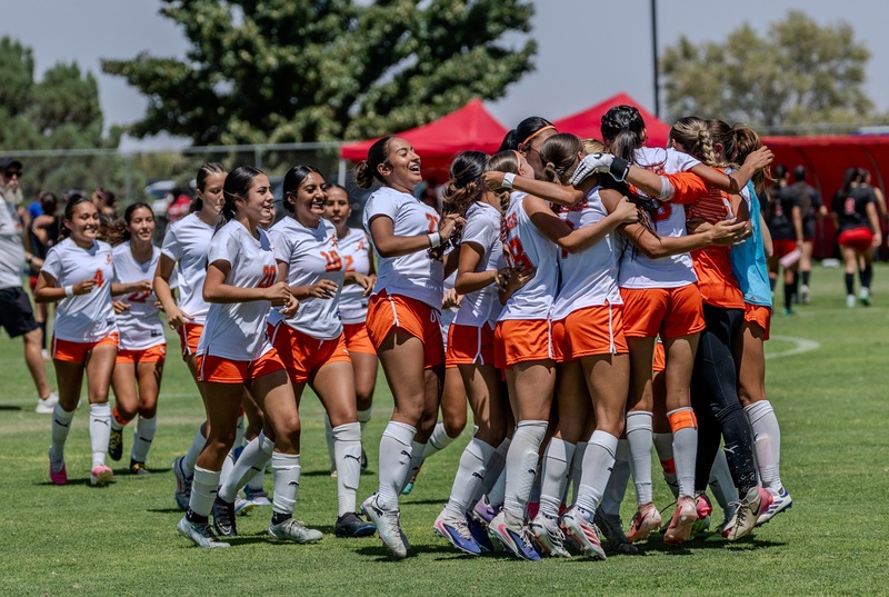 A girls' soccer team in white and orange uniforms jumps, smiles, and hugs one another after winning a game.