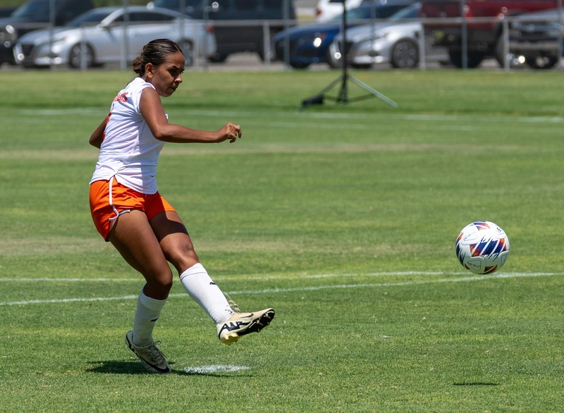 A teen girl in a white and orange soccer uniform takes a penalty kick with her left foot.