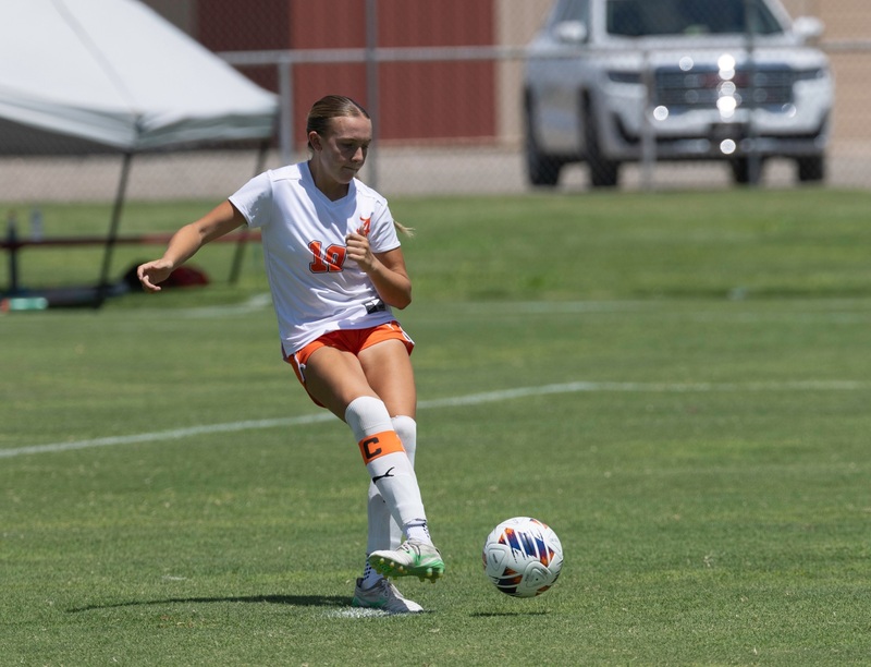 A teen girl in a white and orange soccer uniform takes a penalty kick with her right foot.