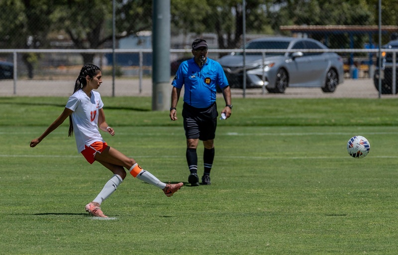 A teen girl in a white and orange soccer uniform takes a penalty kick with her right foot as a referee in blue looks on.