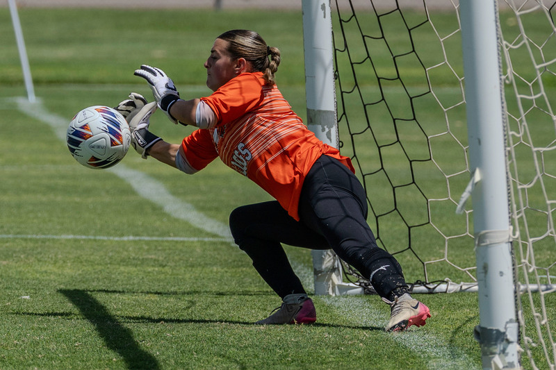 A teen girl in an orange goalie jersey and black pants dives to her right to block a penalty kick.