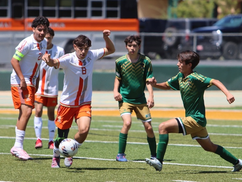A teen boy in a white and orange soccer uniform kicks the ball as two teammates look on and an opponent in green and gold runs in to try to steal the ball.