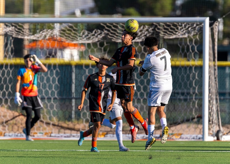 A teen boy in a black and orange soccer uniform heads the ball away from an opponent in white as teammates look on.