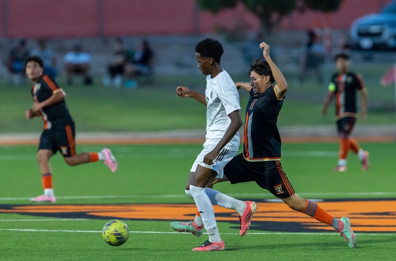 A teen boy in a black and orange soccer uniform lunges to kick the ball with his right foot away from an opponent in white.