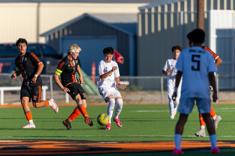 A teen boy in a black and orange soccer uniform taps the ball with his right foot past an opponent in white.