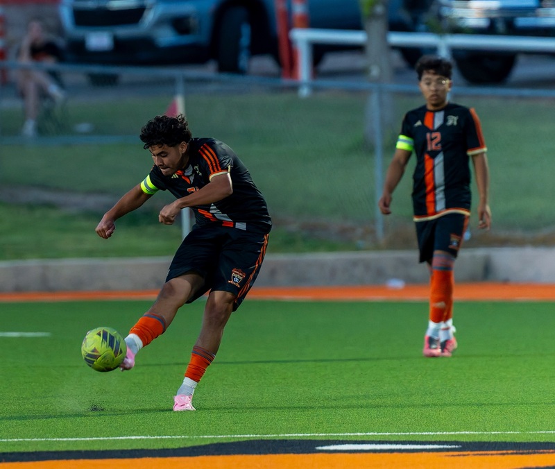 A teen boy in a black and orange soccer uniform kicks the ball as a teammate looks on.