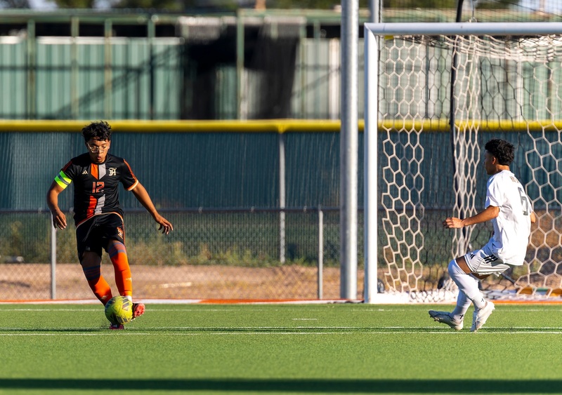 A teen boy in a black and orange soccer uniform kicks the ball as an opponent in white looks on.