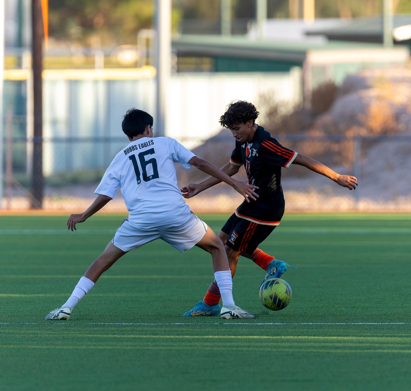 A teen boy in a black and orange soccer uniform taps the ball backward away from an opponent in white.