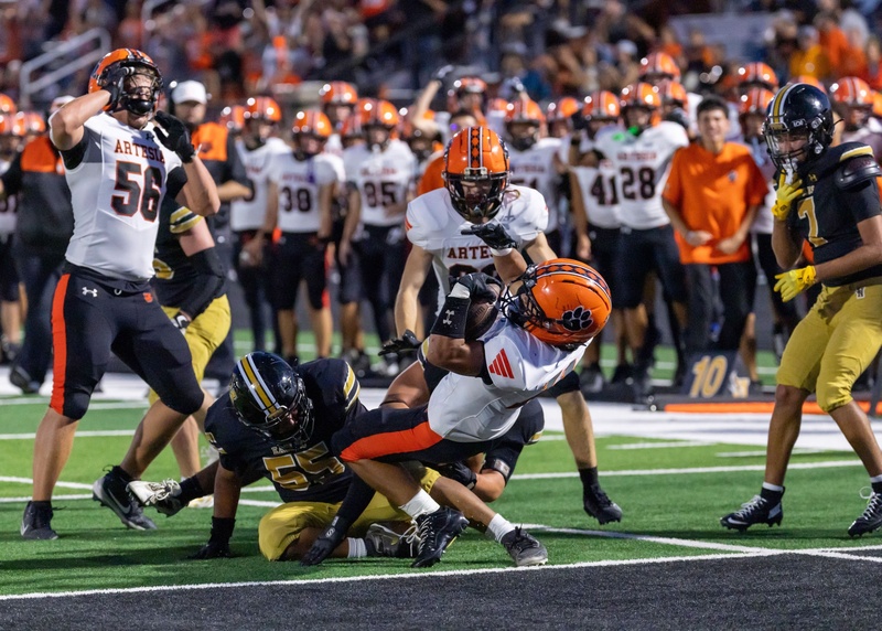 A teen boy in an orange, black and white football uniform dives backward over the goal line with the ball in his arms. A defender in black and gold is hanging from his legs.