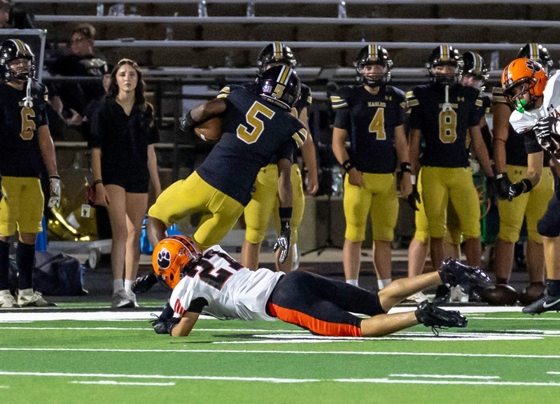 A teen boy in an orange, black and white football uniform trips up an opposing running back in black and gold.