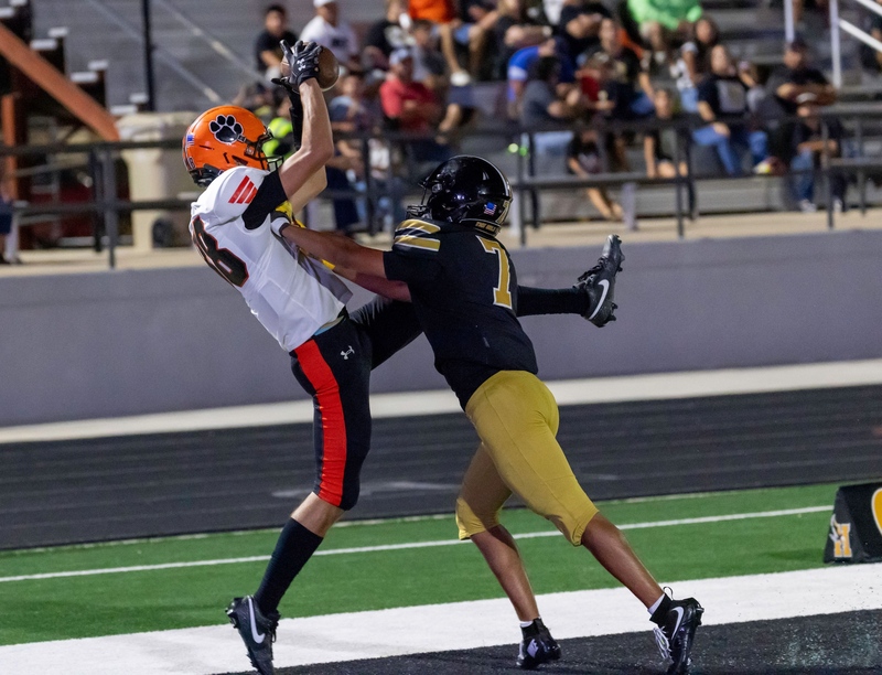 A teen boy in an orange, black and white football uniform leaps into the air to catch a ball over a defender in black and gold.