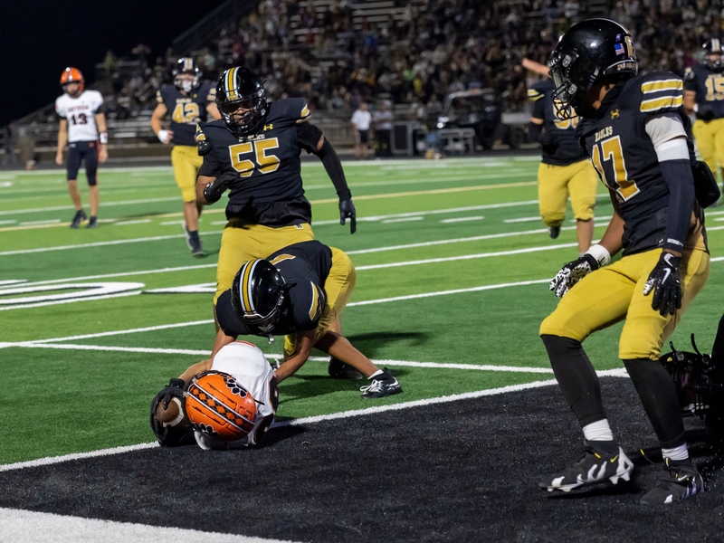 A teen boy in an orange, black and white football uniform lands over the goal line with the ball as a defender in black and gold falls on top of him.