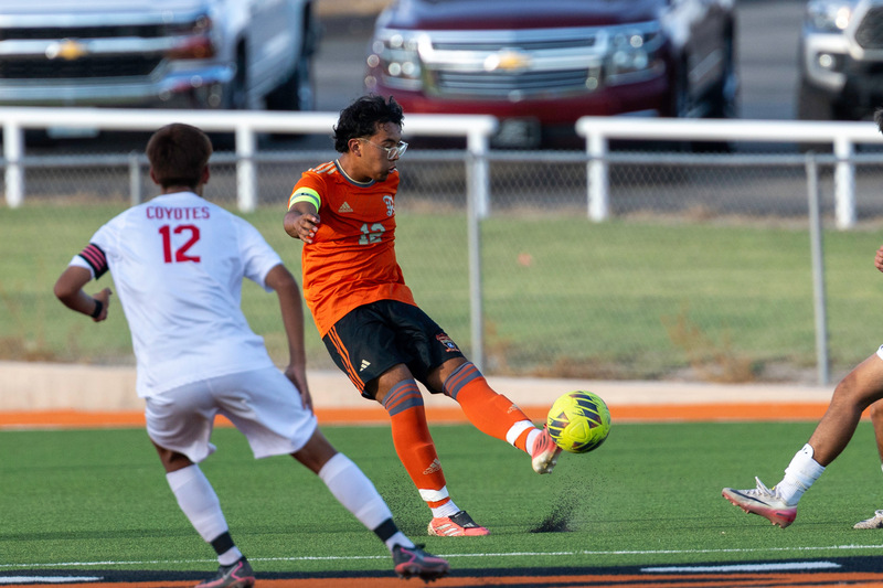 A teen boy in an orange and black soccer uniform kicks a neon-yellow ball as an opponent in white and red looks on.