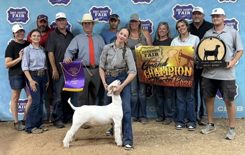 A teen girl holds the head of a white goat. Behind her stand multiple people, three of whom are holding banners.
