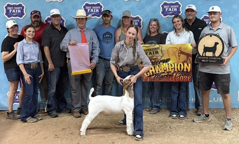 A teen girl holds the head of a white-and-brown goat. Behind her stand multiple people, three of whom are holding banners.