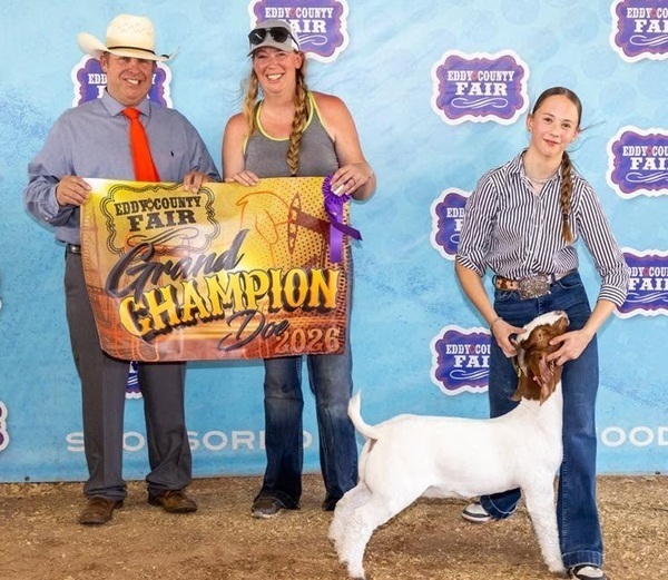 A teen girl holds the head of a white-and-brown goat. Beside her stand a man and a woman holding a banner.