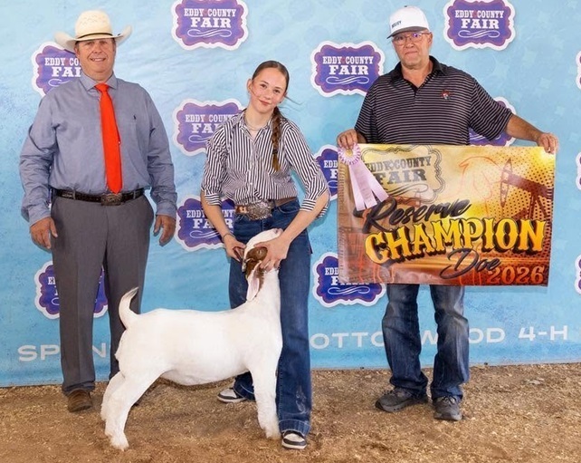 A teen girl holds the head of a white-and-brown goat. On either side of her stand two men, one of whom is holding a banner.