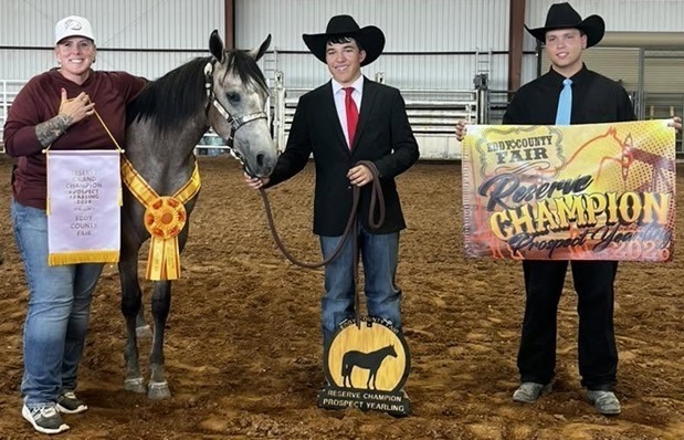 A teen boy in a suit jacket and cowboy hat holds the rein of a grey-and-white horse. Beside him stand a man and a woman, both holding banners.
