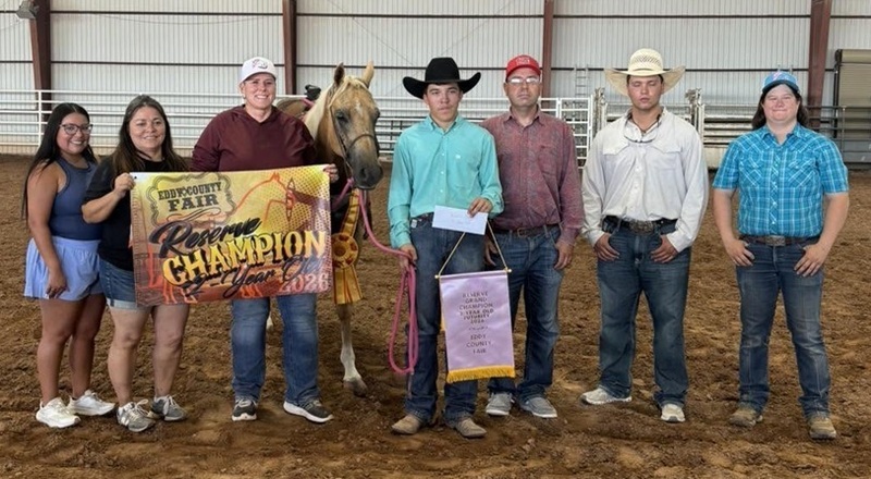 A teen boy in a black cowboy hat holds the rein of a tan horse and a banner. Alongside him stand several people, two of whom are holding a banner.