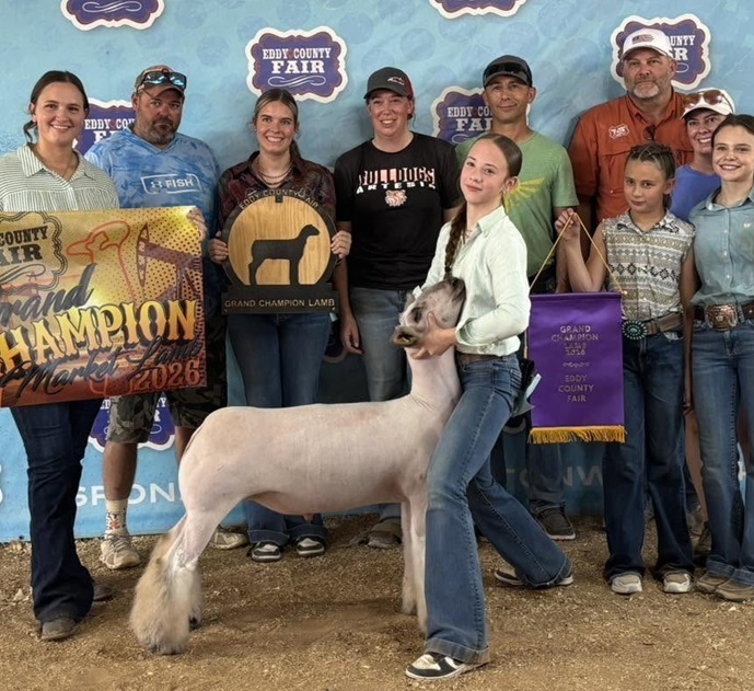 A teen girl holds the head of a shaved sheep. Several people stand behind her, three of whom are holding banners.
