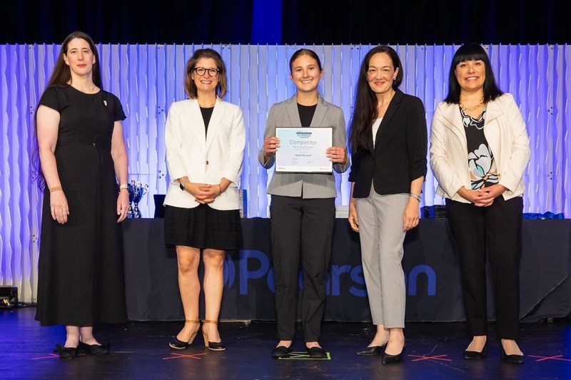A teen girl in a black-and-grey pant suit holds a certificate, surrounded by four women.