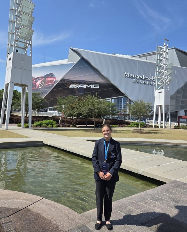 A teen girl in a pant suit stands in front of a water feature in front of a large building reading "Mercedes-Benz Stadium".