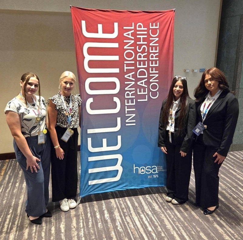 Four teen girls in business-casual attire stand alongside a banner reading "Welcome International Leadership Conference".