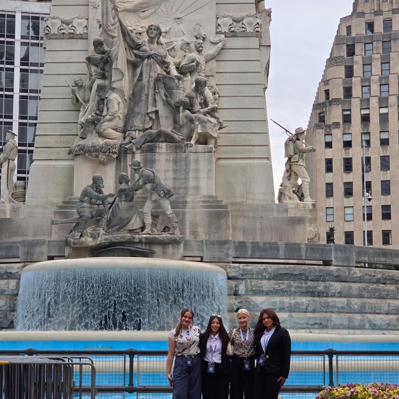 Four teen girls in business-casual attire stand in front of a large, ornate fountain in front of a building.