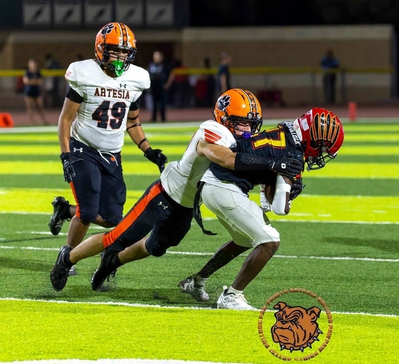 A teen boy in an orange, white and black football uniform lunges to tackle an opponent in black, red and gold who is carrying the ball.