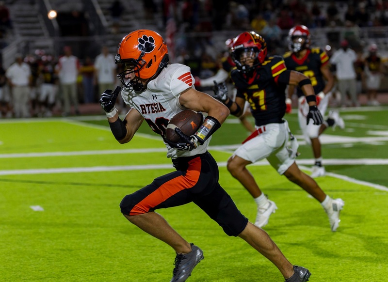 A teen boy in an orange, white and black football uniform runs down the sideline with the ball tucked into his left elbow. Opponents in black, red and gold chase him.