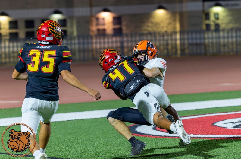 A teen boy in an orange, white and black football uniform falls into his back in the end zone while clutching the ball. One opponent in black, red and gold is landing atop him as another runs into the frame.