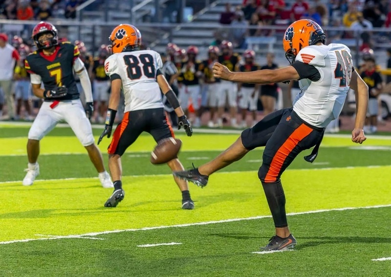 A teen boy in an orange, white and black football uniform punts the ball. In the foreground are a teammate and an opponent in black, red and gold.