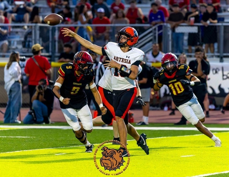 A teen boy in an orange, white and black football uniform releases the ball from his right hand as two opponents in black, red and gold close in from behind.