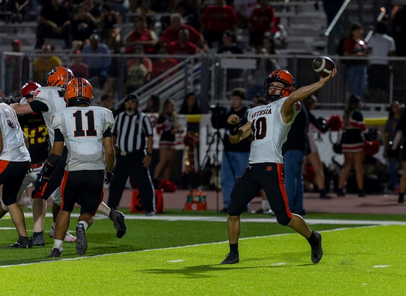 A teen boy in an orange, white and black football uniform prepares to release the ball from his left hand.