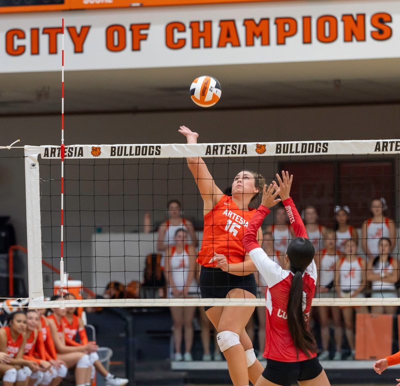 A teen girl in an orange and black volleyball uniform spikes an orange, white and black ball over the net as an opponent in red and white puts her hands up.
