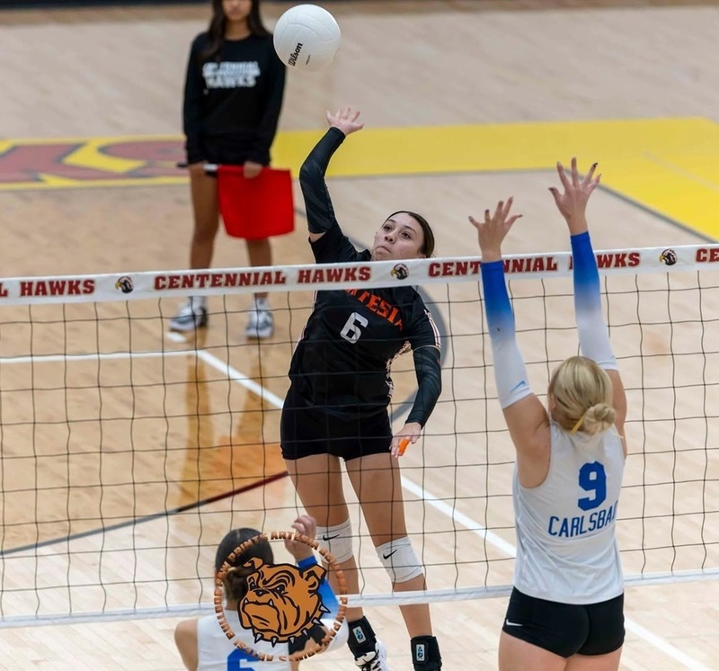 A teen girl in a black volleyball uniform prepares to spike the ball as an opponent in blue and white jumps up with her hands in the air to block.