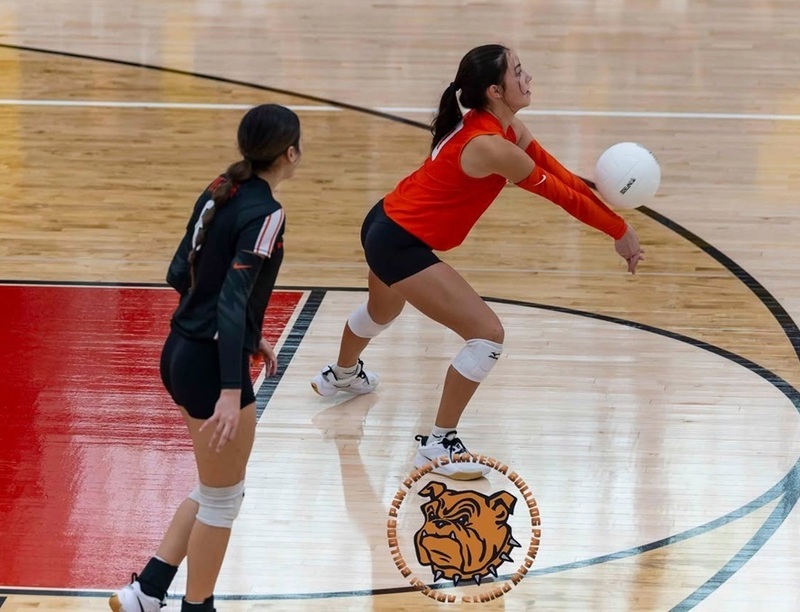 A teen girl in an orange and black volleyball uniform bump-passes the ball from the back row.