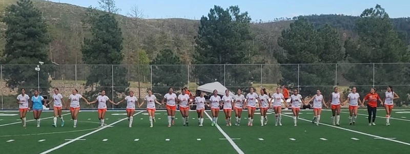 A high school girls' soccer team in white and orange uniforms joins hands as they walk across a field toward the camera. Behind them, pine trees and hills can be seen.