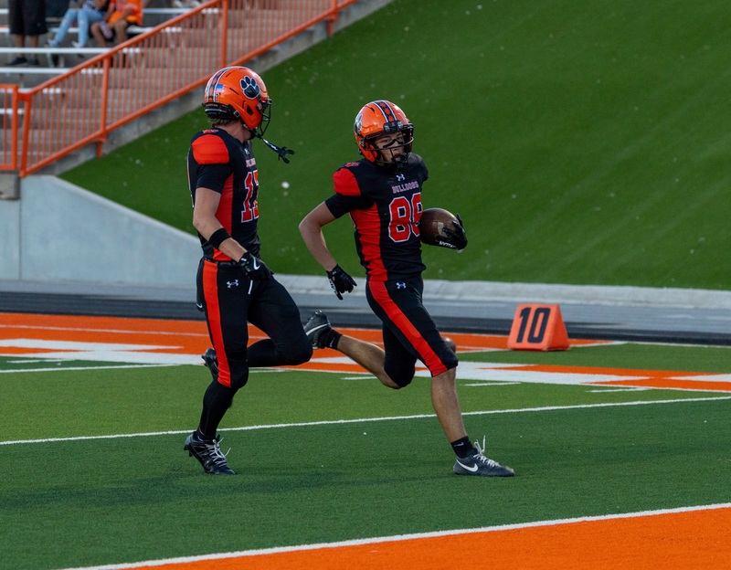 A teen boy in an orange and black football uniform carries the ball toward the goal line in his left elbow as a teammate runs alongside him.