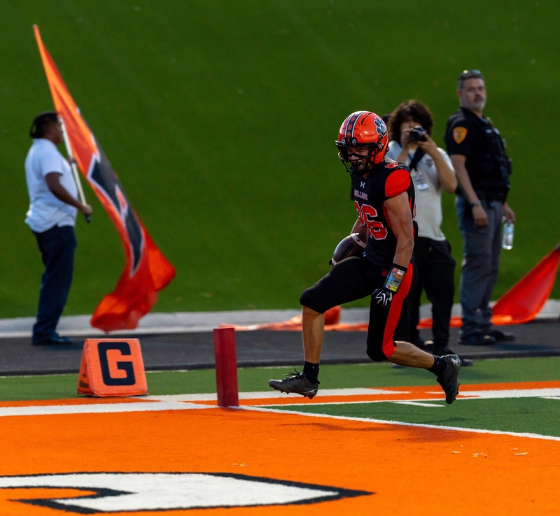 A teen boy in an orange and black football uniform carries the ball in his right hand as he steps over the goal line.